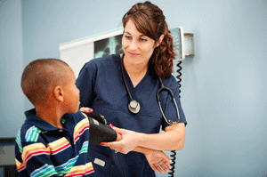 A female health care provider is smiling at a young patient and placing a blood pressure cuff on his arm.