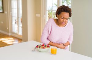 Woman in pink sweater experiencing abdominal pain while eating breakfast