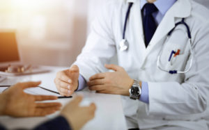 A doctor is talking with his patient while they are seated together at a desk in a sunny office. The physician is recording the patient's medical history on a clipboard.