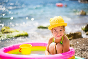 Little girl in a yellow straw hat playing with a bucket in an inflatable pool on the beach