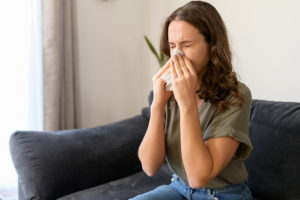 Young woman sneezing into tissue at home, feeling unwell from seasonal allergies