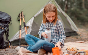 Young blonde girl drinking water and sitting near her tent while camping in a forest