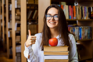 Smiling female college student giving thumbs-up with stack of boxes and apple